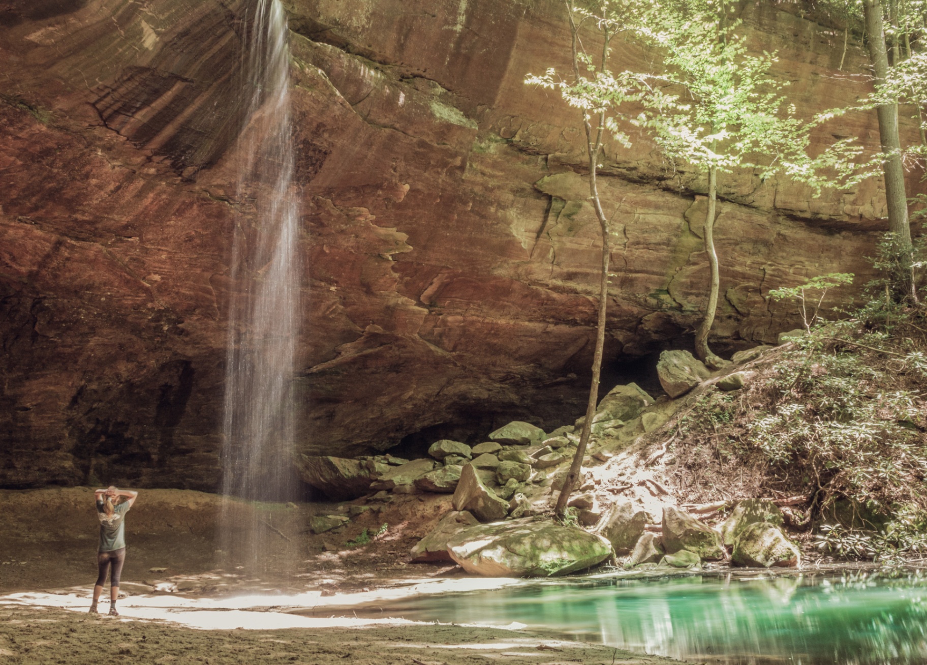 Hiker beneath a tall waterfall in a sandstone alcove at Red River Gorge