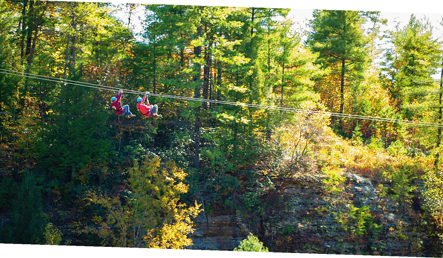 Two zipline riders side by side over fall foliage at Red River Gorge